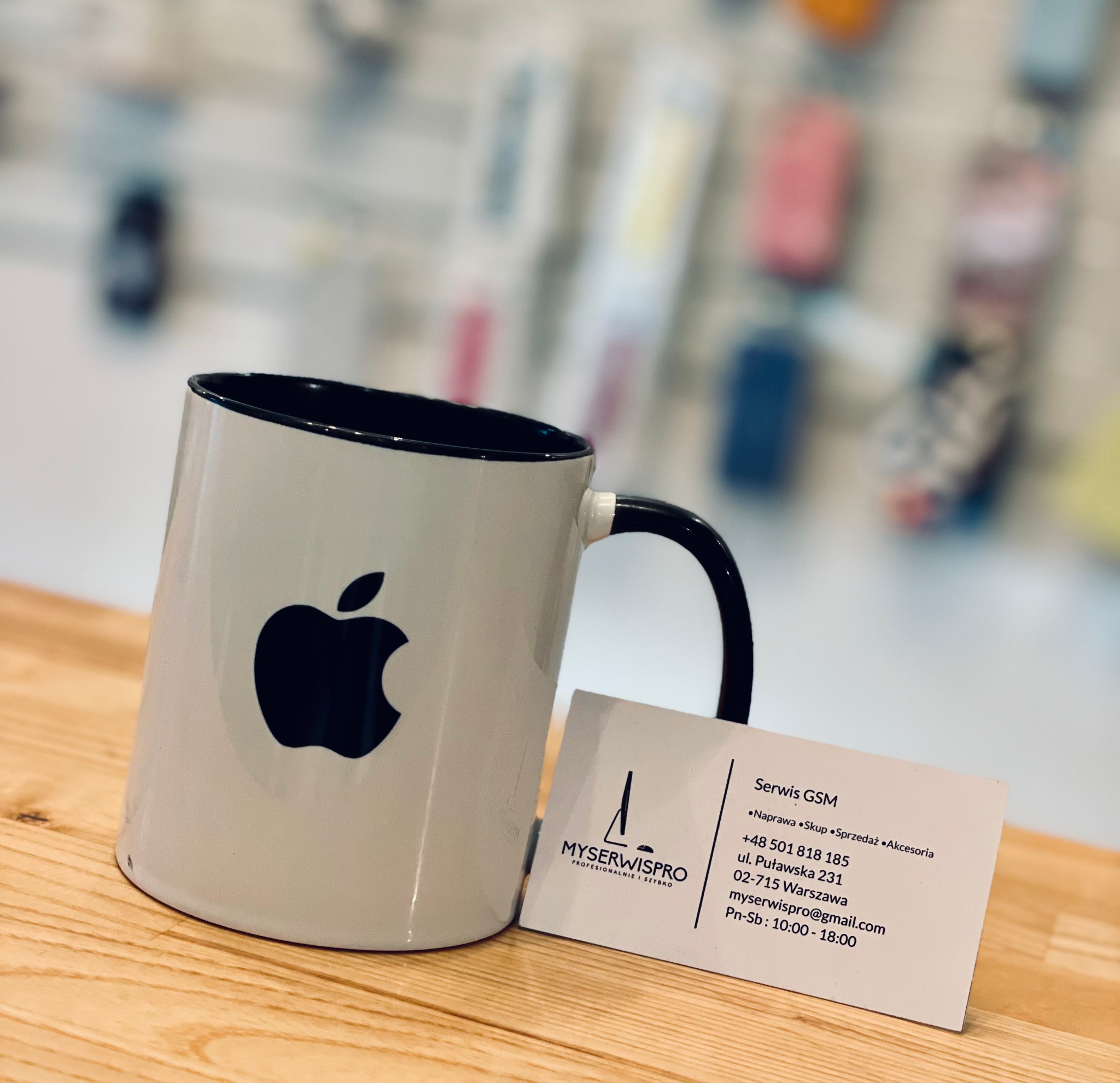 Kubek z logo Apple i wizytówka serwisu GSM na drewnianym blacie, w tle rozmyte akcesoria telefoniczne.