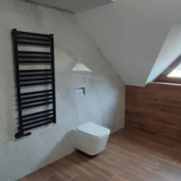 Wykończona łazienka na poddaszu ze skośnym sufitem, brązowe płytki imitujące drewno na podłodze i częściowo na ścianie, wisząca toaleta, czarny grzejnik drabinkowy na szarej ścianie.
