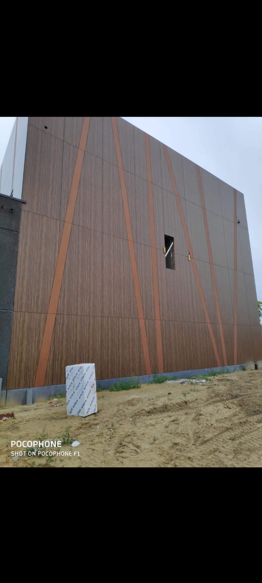 Elewacja budynku wykończona panelami imitującymi drewno, z dekoracyjnymi, ukośnymi pasami w kolorze miedzianym, widoczny fragment placu budowy i zapakowane panele elewacyjne.