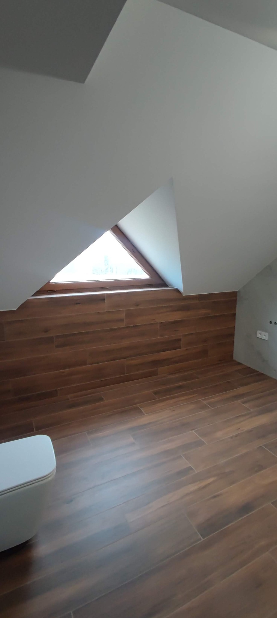 Poddasze po remoncie z drewnopodobną okładziną ścienną i podłogową, trójkątne okno w skosie dachu, fragment białej toalety w rogu pomieszczenia.