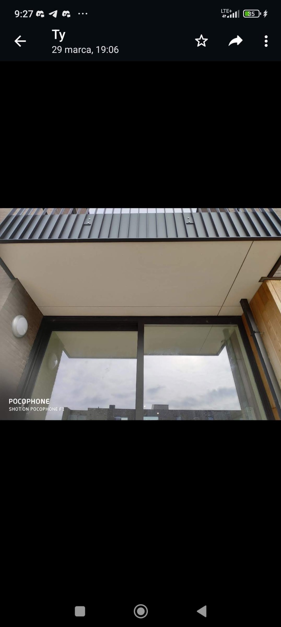 Widok z dołu na balkon z metalową balustradą i dużym oknem, widoczne elementy wykończenia elewacji budynku.