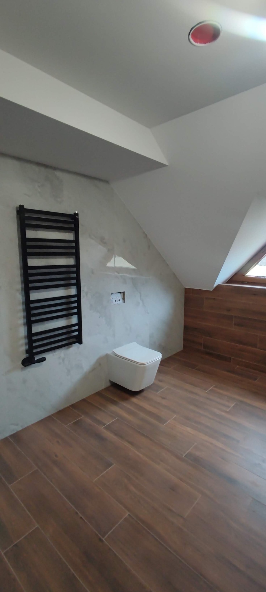 Wykończona łazienka na poddaszu ze skośnym sufitem, brązowe płytki imitujące drewno na podłodze i częściowo na ścianie, wisząca toaleta, czarny grzejnik drabinkowy na szarej ścianie.