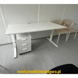 Białe biurko z regulowaną wysokością, trzema szufladami i dwoma krzesłami w tle, ustawione w minimalistycznym biurze z panelami ściennymi.