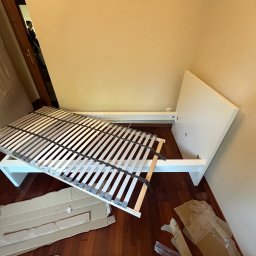 Montaż mebli Zielona Góra - Biała rama łóżka z listwami w trakcie montażu, kartony i folie na parkiecie, widoczny fragment buta osoby montującej.