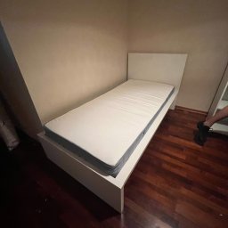 Montaż mebli Zielona Góra - Pojedyncze białe łóżko z materacem w rogu pokoju, widoczne ciemne drewniane panele podłogowe i fragment nogi osoby stojącej obok.