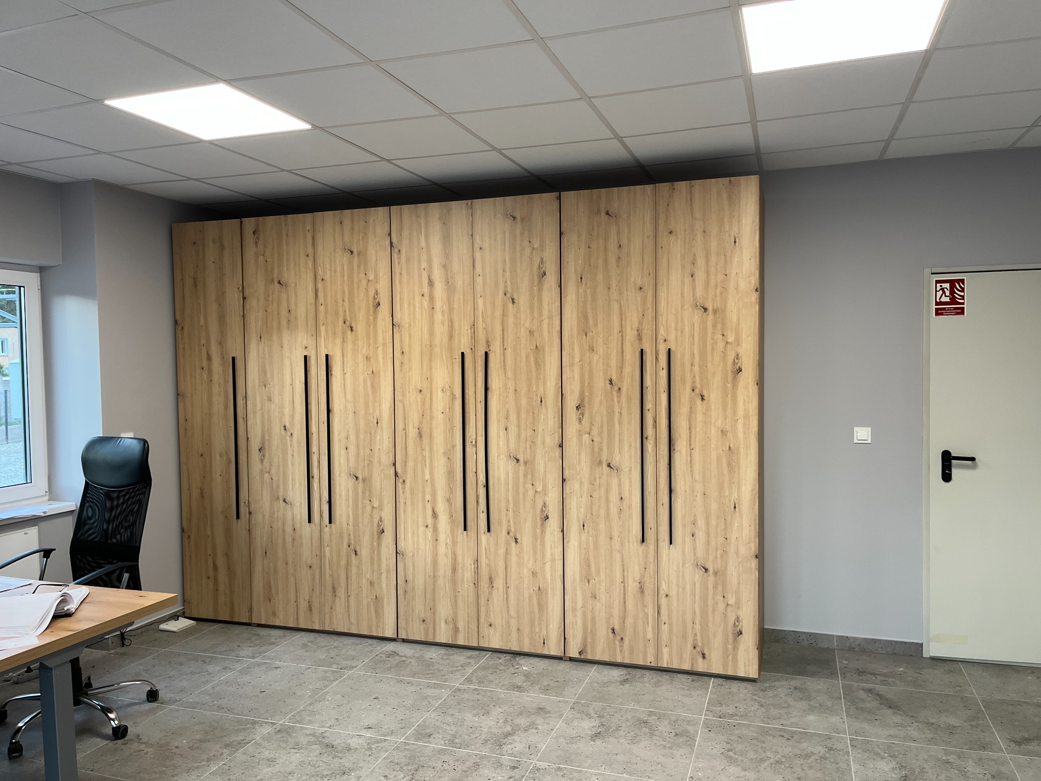 Duża, drewniana szafa z czarnymi uchwytami w biurze, widok na szafę, biurko z krzesłem i drzwi ewakuacyjne.