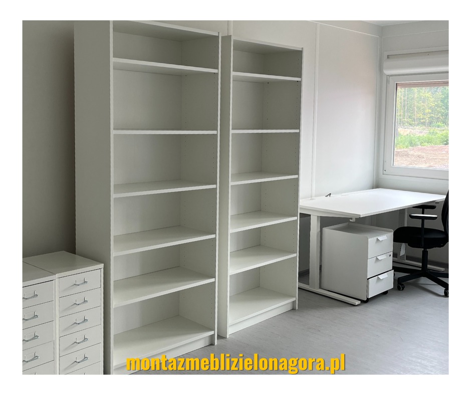 Jasne biuro z dwoma wysokimi regałami, biurkiem z regulowaną wysokością i kontenerkiem z trzema szufladami, widok przez okno na zieleń.