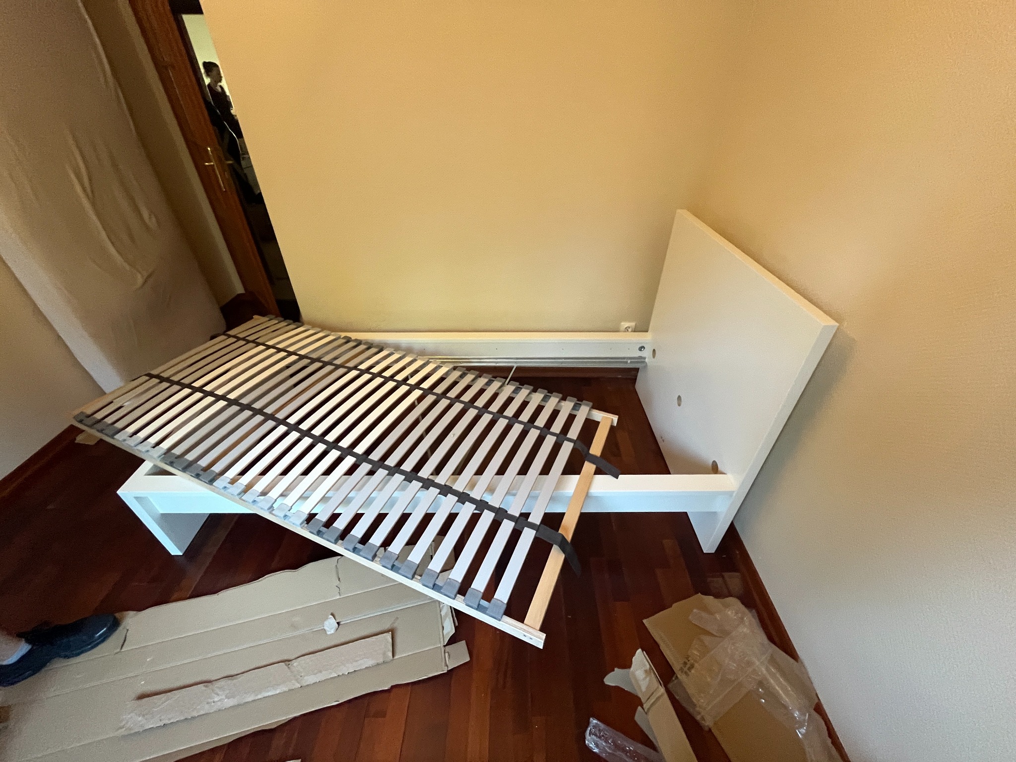 Biała rama łóżka z listwami w trakcie montażu, kartony i folie na parkiecie, widoczny fragment buta osoby montującej.