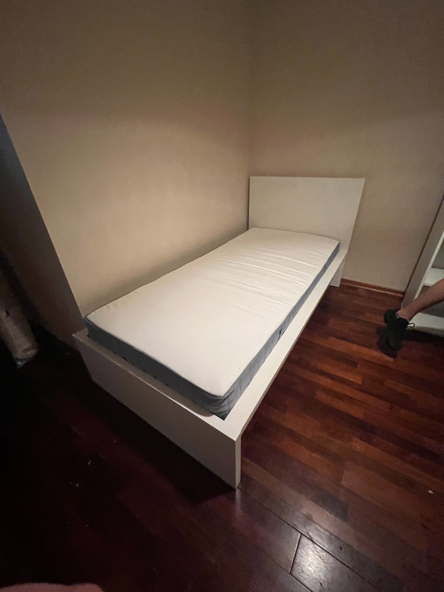 Pojedyncze białe łóżko z materacem w rogu pokoju, widoczne ciemne drewniane panele podłogowe i fragment nogi osoby stojącej obok.