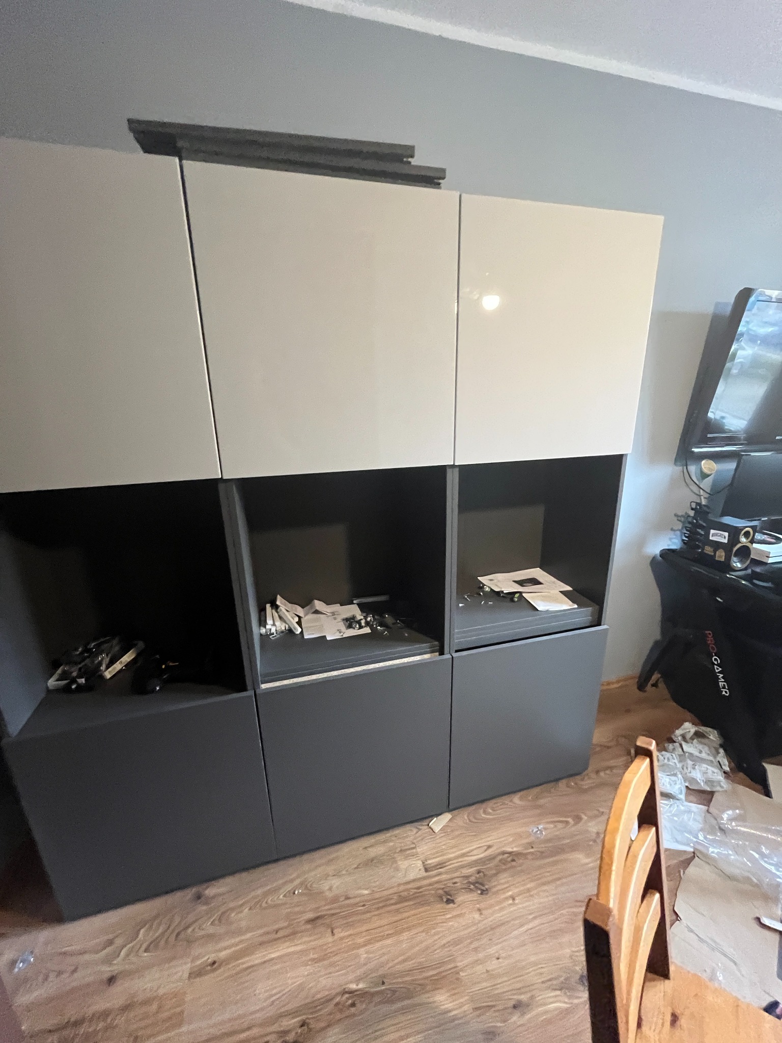 Szafka modułowa w trakcie montażu, widoczne elementy składowe w otwartych półkach, na podłodze resztki opakowań, krzesło w tle.
