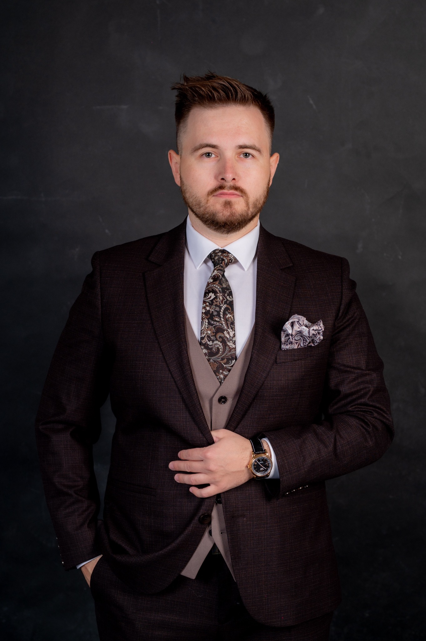 Portret mężczyzny w brązowym garniturze w kratę, z wzorzystym krawatem i poszetką, pozującego na ciemnym tle.