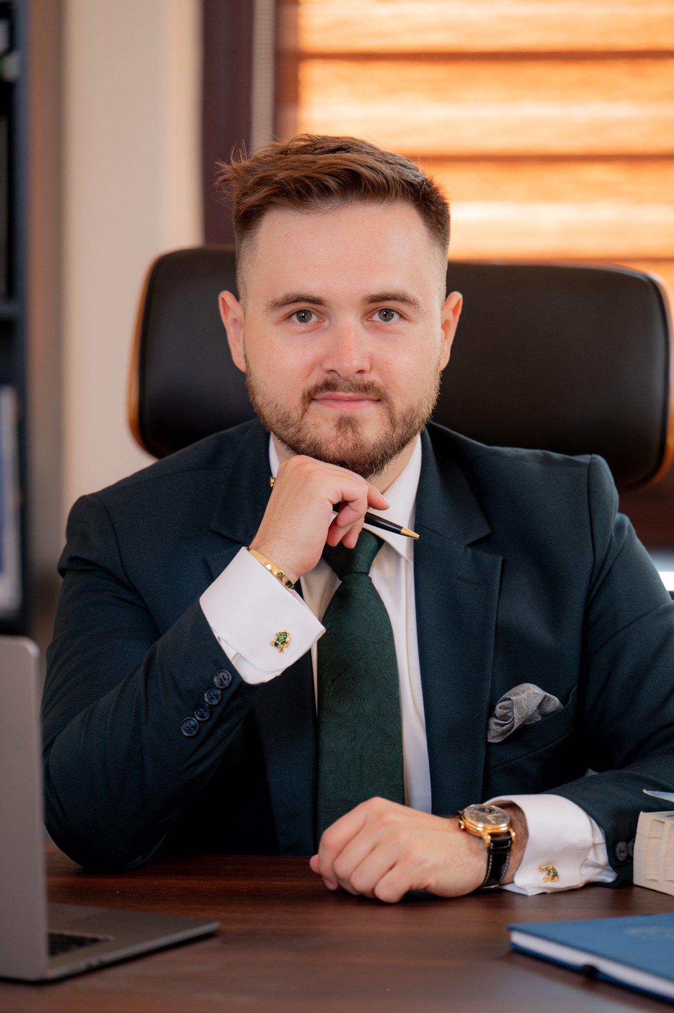 Portret prawnika w ciemnozielonym garniturze, siedzącego za biurkiem z laptopem, trzymającego długopis przy brodzie, z widocznymi złotymi spinkami do mankietów i zegarkiem.