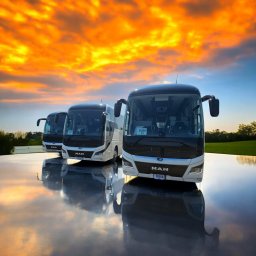 M CHOICE MARCIN MAZUROWSKI - Trzy nowoczesne autobusy MAN ustawione w rzędzie na błyszczącej powierzchni odbijającej zachodzące słońce, tworzące lustrzane odbicie pojazdów i dramatyczne, pomarańczowe niebo.