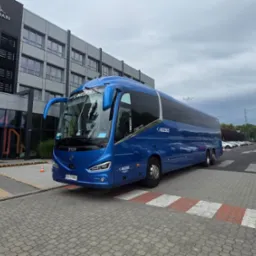 Niebieski autokar marki Irizar parkuje przed budynkiem Saint-Gobain. Widoczny numer rejestracyjny KN 2188E oraz logo BuzzBus. W tle zaparkowane samochody.