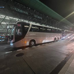 M CHOICE MARCIN MAZUROWSKI - Biały autobus BuzzBus na lotnisku nocą, z otwartymi drzwiami, pasażerowie wsiadają. Widoczne światła i szklana fasada terminalu.