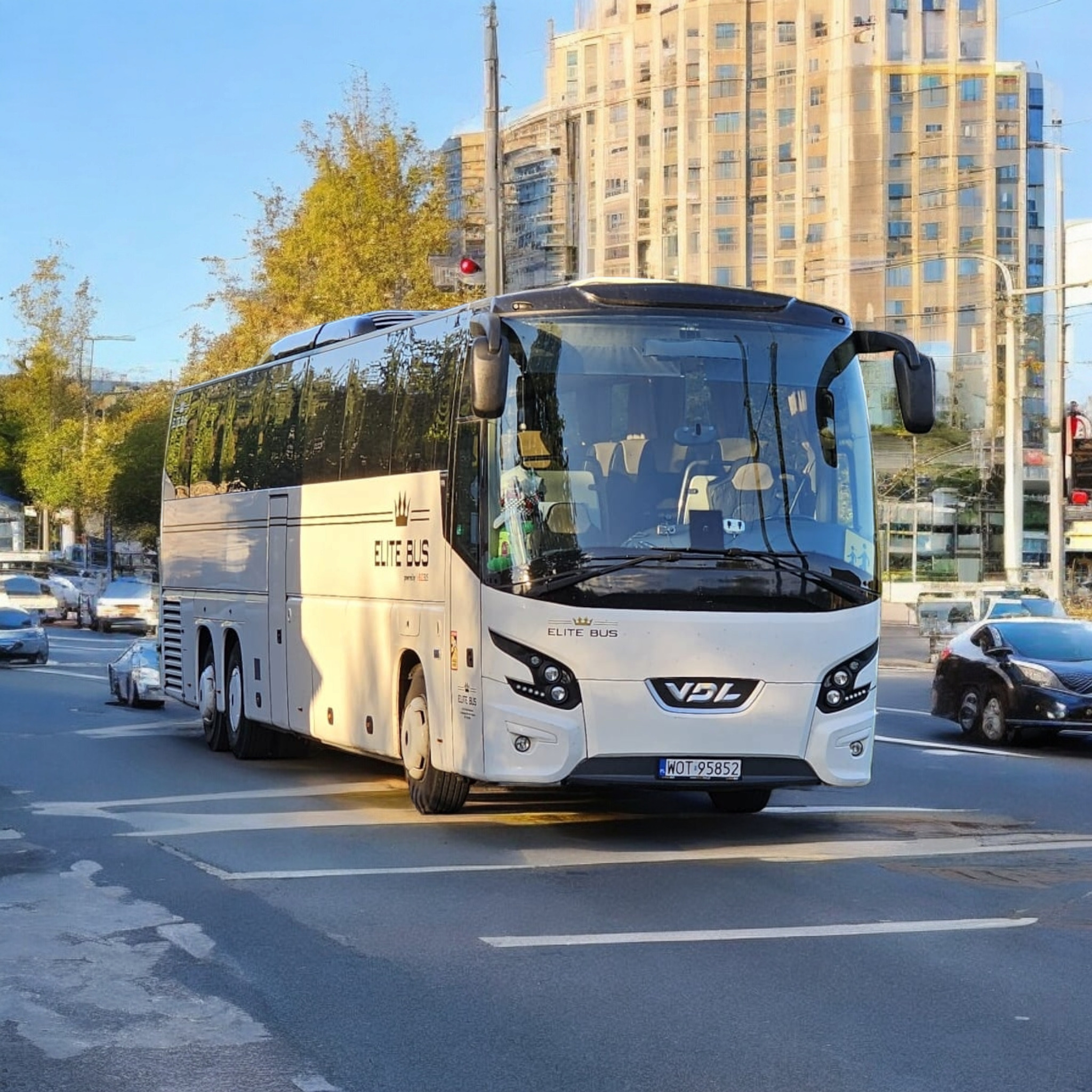 Biały autokar Elite Bus VDL z warszawską rejestracją WOT-95852 przejeżdża przez skrzyżowanie na tle nowoczesnego budynku.
