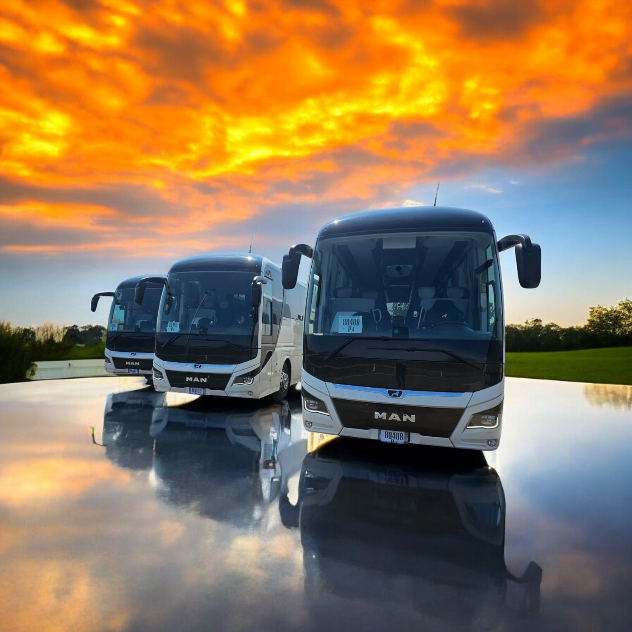 Trzy nowoczesne autobusy MAN ustawione w rzędzie na błyszczącej powierzchni odbijającej zachodzące słońce, tworzące lustrzane odbicie pojazdów i dramatyczne, pomarańczowe niebo.
