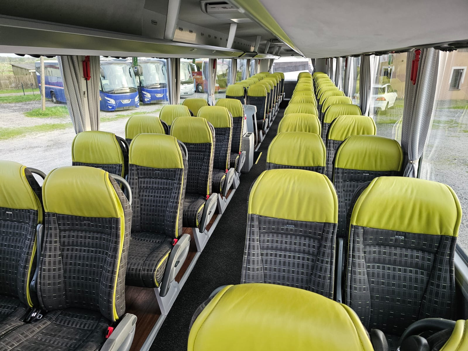 Wnętrze autobusu turystycznego z rzędami foteli z zielonymi zagłówkami i szarą tapicerką w kratę; widok z przodu na puste siedzenia i okna z zasłonami.