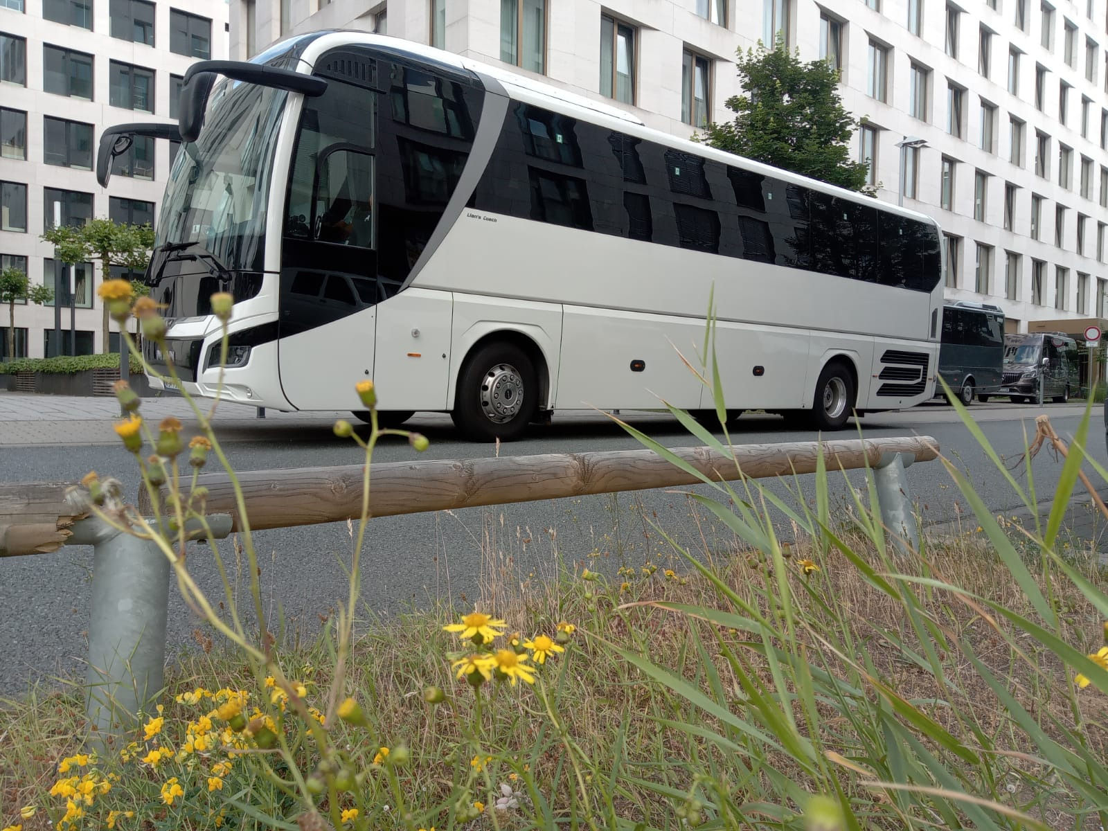Nowoczesny, biały autokar z czarnymi akcentami, zaparkowany w Katowicach na tle biurowca, z widocznymi z przodu żółtymi kwiatami i drewnianą barierką.