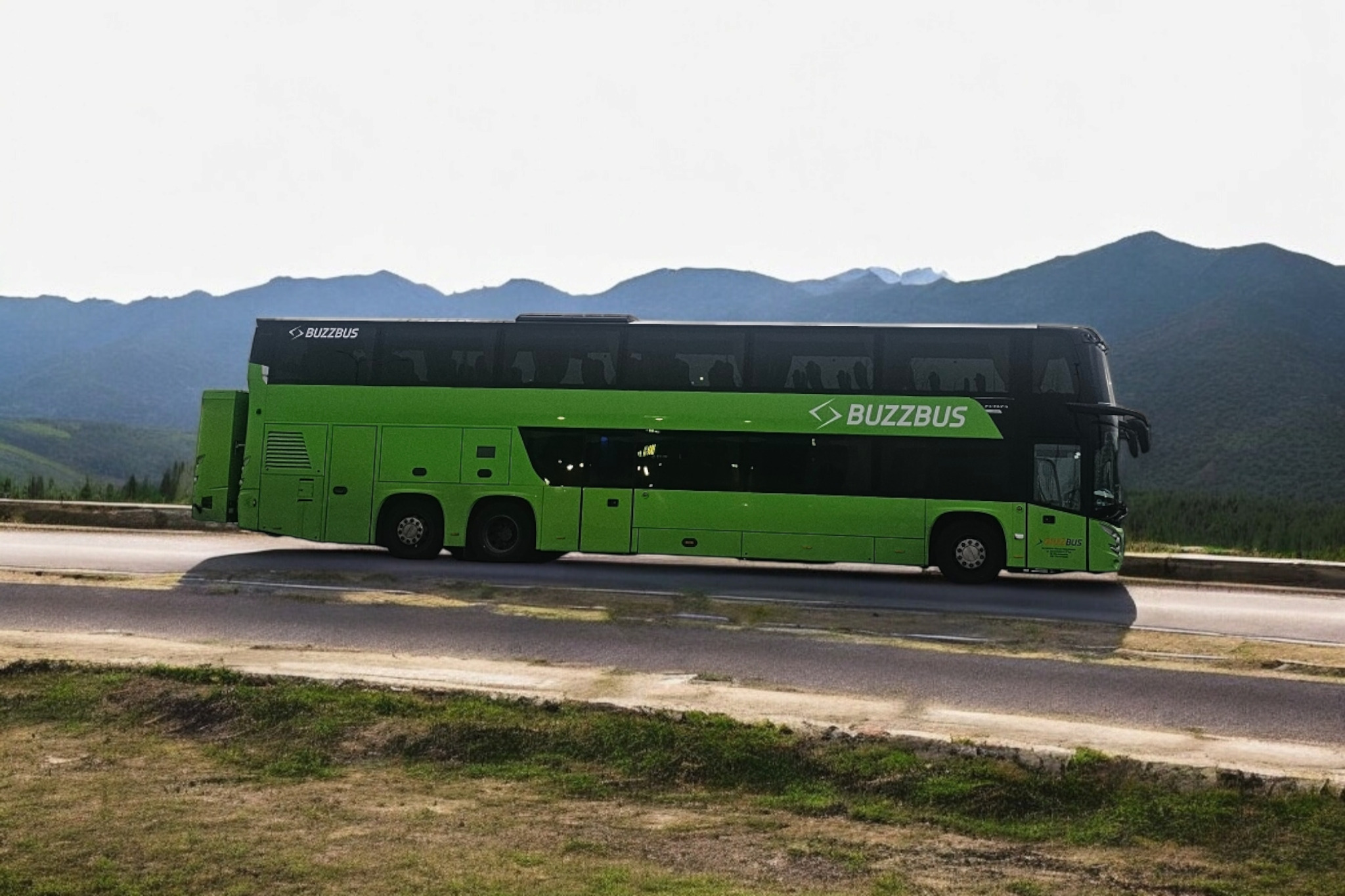 Zielony, dwupiętrowy autokar 'Buzzbus' na drodze z górami w tle, widok z boku.