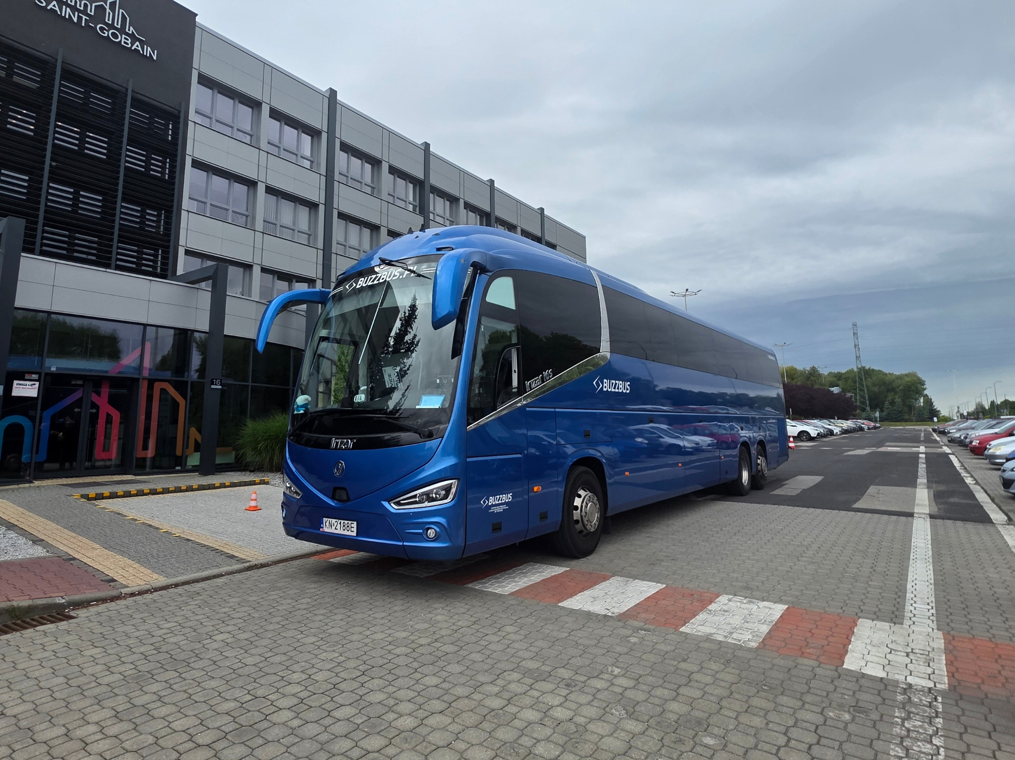 Niebieski autokar marki Irizar parkuje przed budynkiem Saint-Gobain. Widoczny numer rejestracyjny KN 2188E oraz logo BuzzBus. W tle zaparkowane samochody.