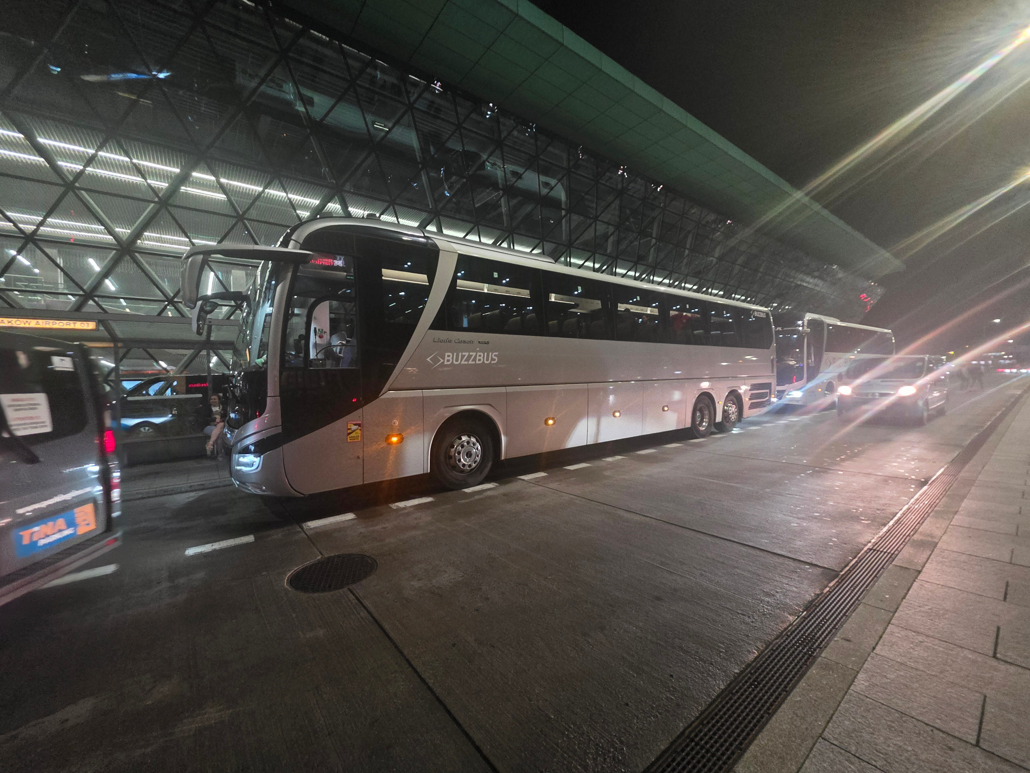Biały autobus BuzzBus na lotnisku nocą, z otwartymi drzwiami, pasażerowie wsiadają. Widoczne światła i szklana fasada terminalu.
