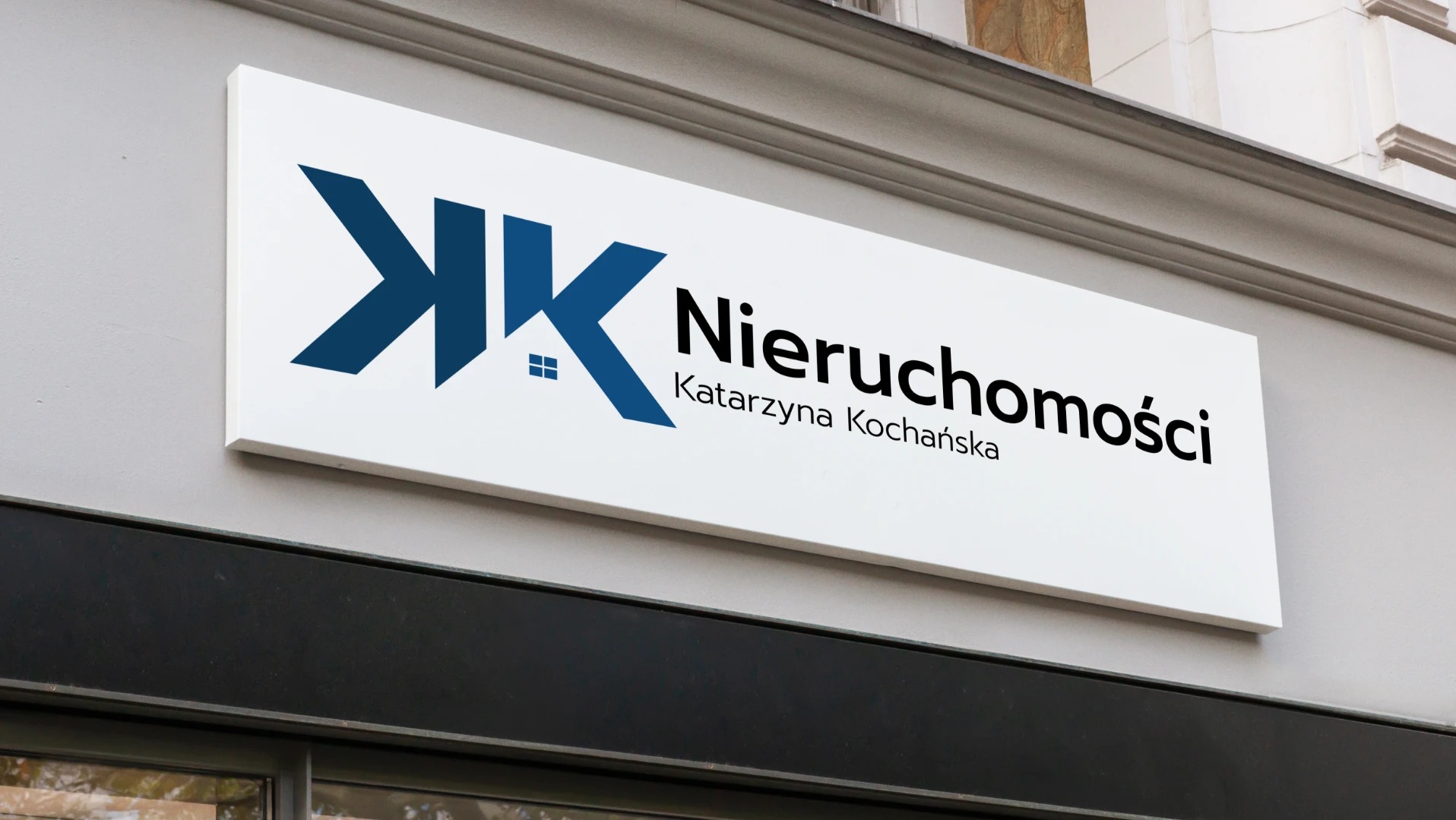 Szyld reklamowy z niebieskim logo nieruchomości, zawierającym stylizowany dom wkomponowany w litery 'KK' i czarny napis 'Nieruchomości Katarzyna Kochańska' na białym tle, zamontowany nad wejściem...