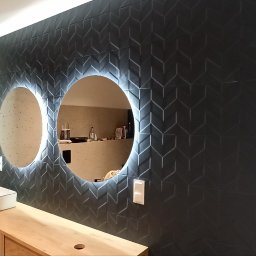 Glazurex - Nowoczesna łazienka z czarnymi płytkami w geometryczny wzór, podświetlanymi okrągłymi lustrami, umywalką nablatową i drewnianą szafką pod umywalką.