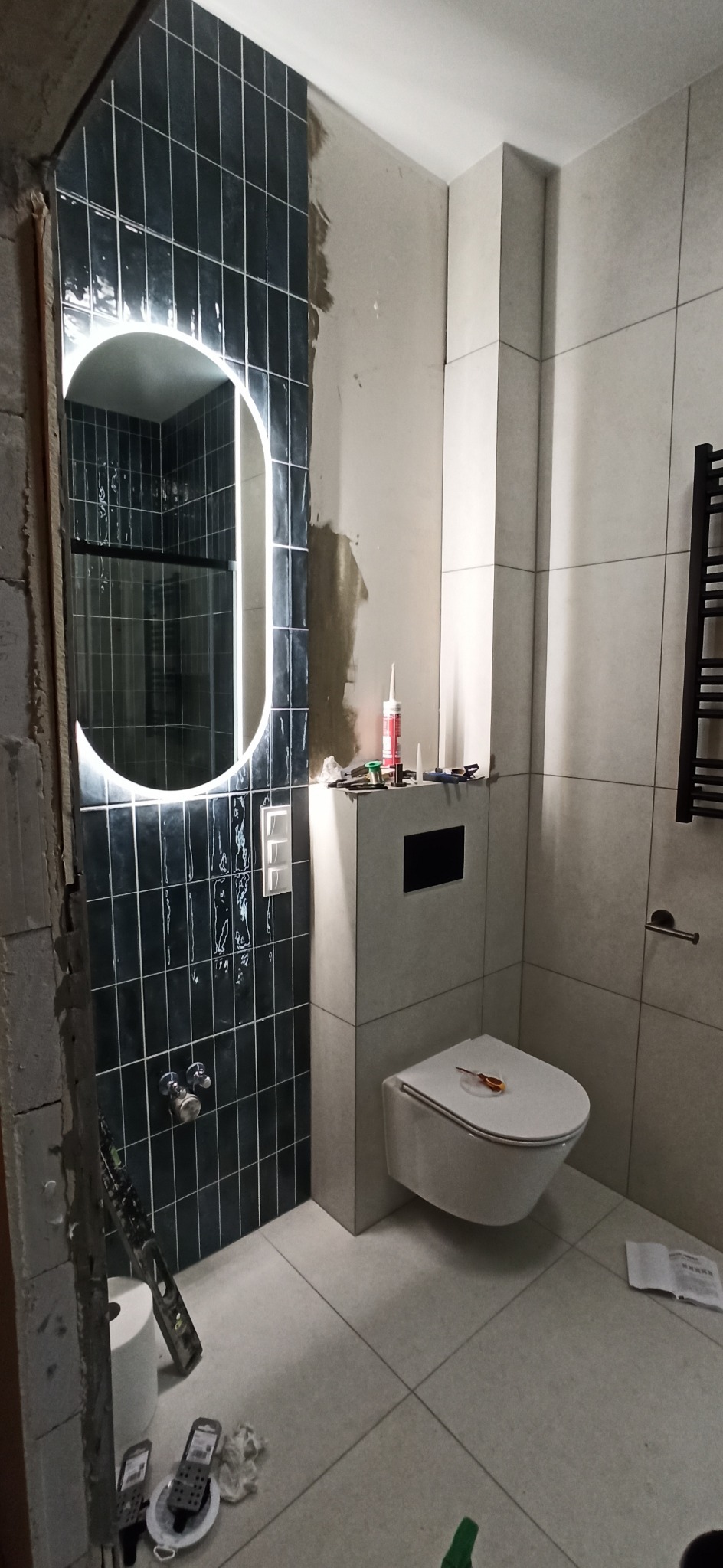 Remont łazienki w trakcie: czarne płytki z białą fugą, podświetlane owalne lustro, biała wisząca toaleta z deską, narzędzia i materiały budowlane widoczne na podłodze i obudowie stelaża podtynkowego.