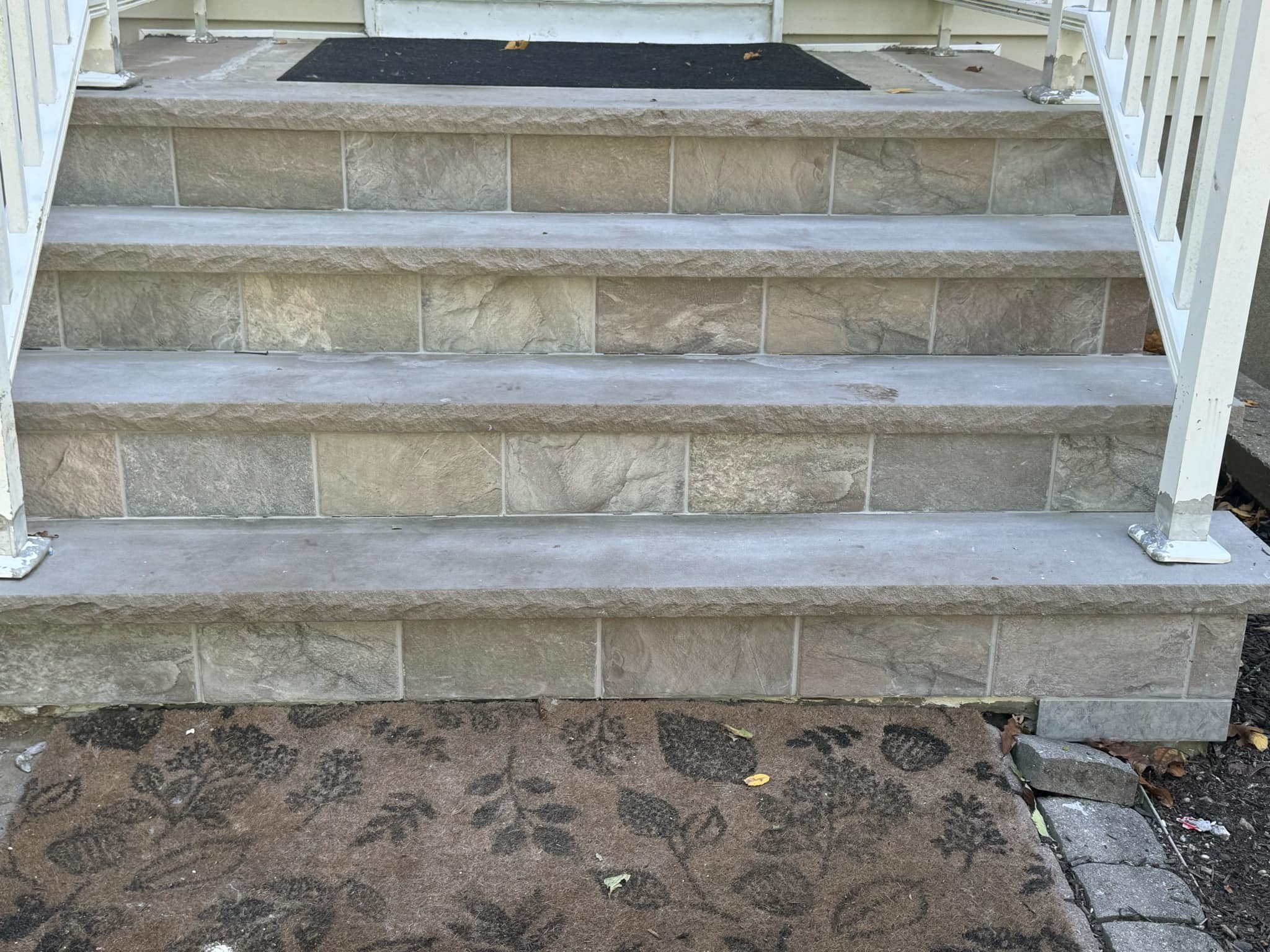 Schody wejściowe wyłożone szarymi płytkami imitującymi kamień, z białą metalową balustradą, widok z dołu na wycieraczkę z motywem roślinnym.