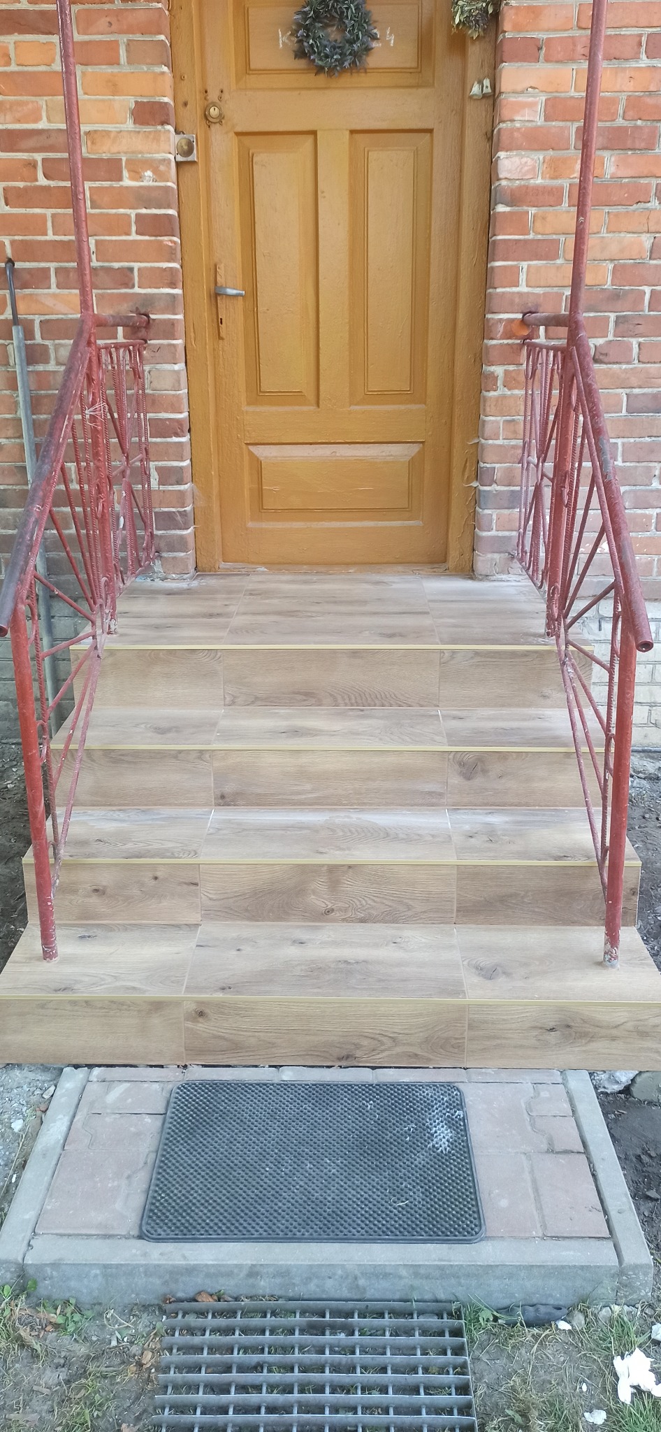 Wyłożone płytkami imitującymi drewno schody wejściowe do budynku z cegły, z pomalowaną na żółto drewnianą stolarką drzwiową i czerwoną metalową barierką.