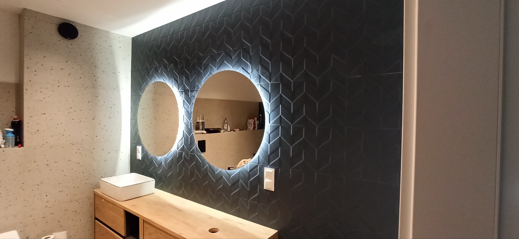 Nowoczesna łazienka z czarnymi płytkami w geometryczny wzór, podświetlanymi okrągłymi lustrami, umywalką nablatową i drewnianą szafką pod umywalką.