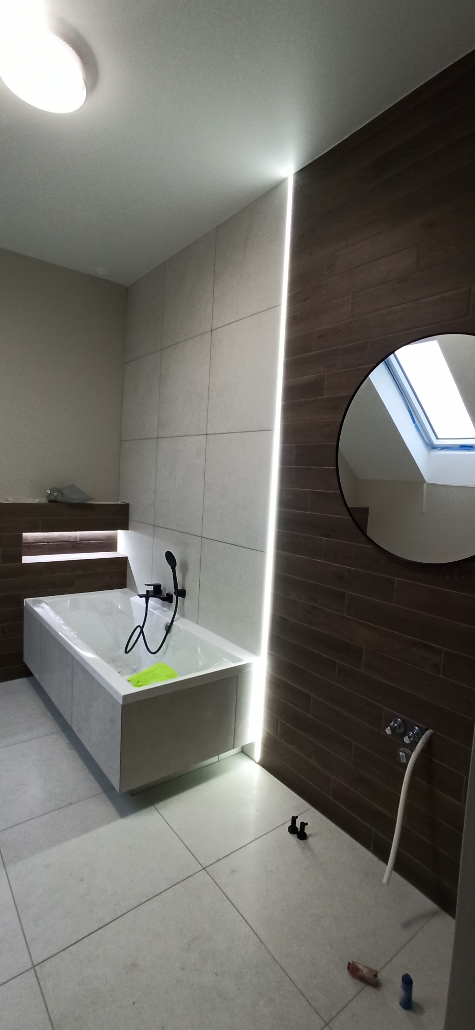 Nowoczesna łazienka z podświetlaną wanną, brązowymi płytkami imitującymi drewno i okrągłym lustrem odbijającym okno dachowe.