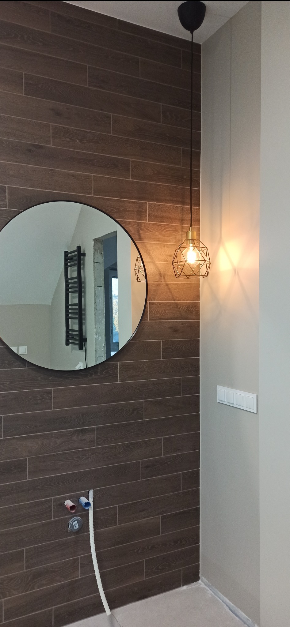 Łazienka w trakcie wykańczania z brązowymi płytkami imitującymi drewno, okrągłe lustro z widocznym grzejnikiem drabinkowym w odbiciu, wisząca lampa z metalowym kloszem i widocznymi przyłączami...