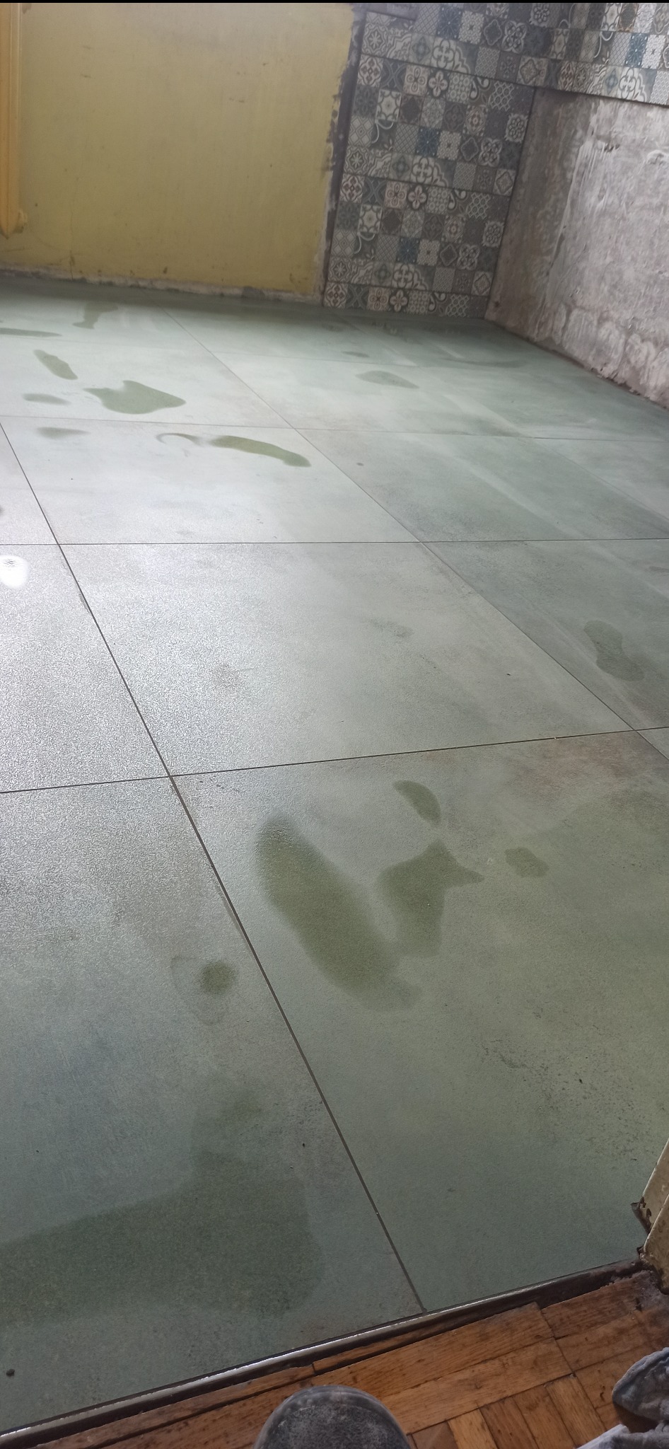 Świeżo położone, jasnoszare płytki podłogowe w trakcie schnięcia, z widocznymi mokrymi śladami stóp i smugami wody, fragment ściany z dekoracyjnymi płytkami i fragment żółtej ściany w tle.