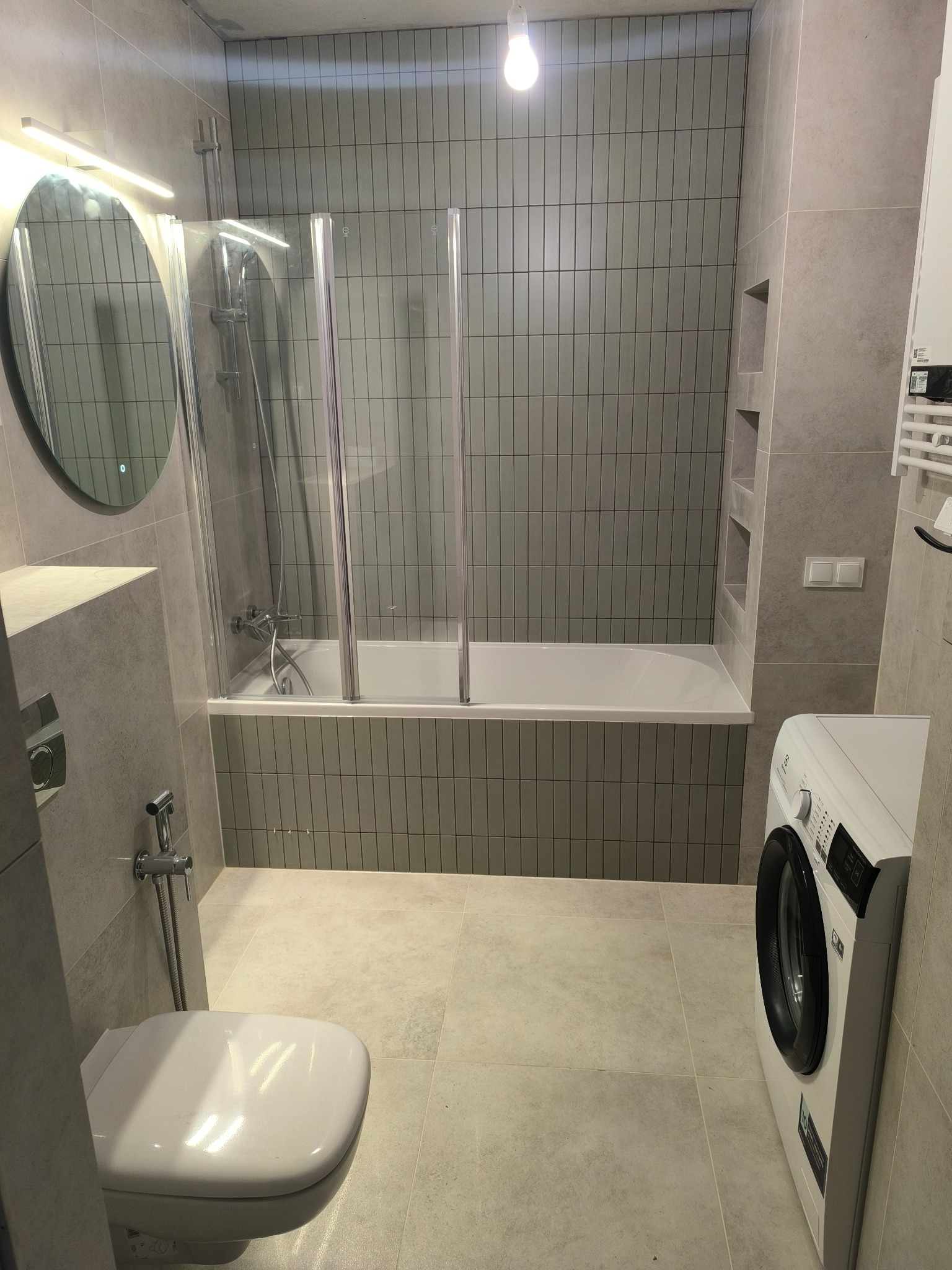 Nowoczesna łazienka w odcieniach szarości z pionowymi płytkami, kabiną nawannową, okrągłym lustrem z podświetleniem LED, białą toaletą z bidetką i pralką.