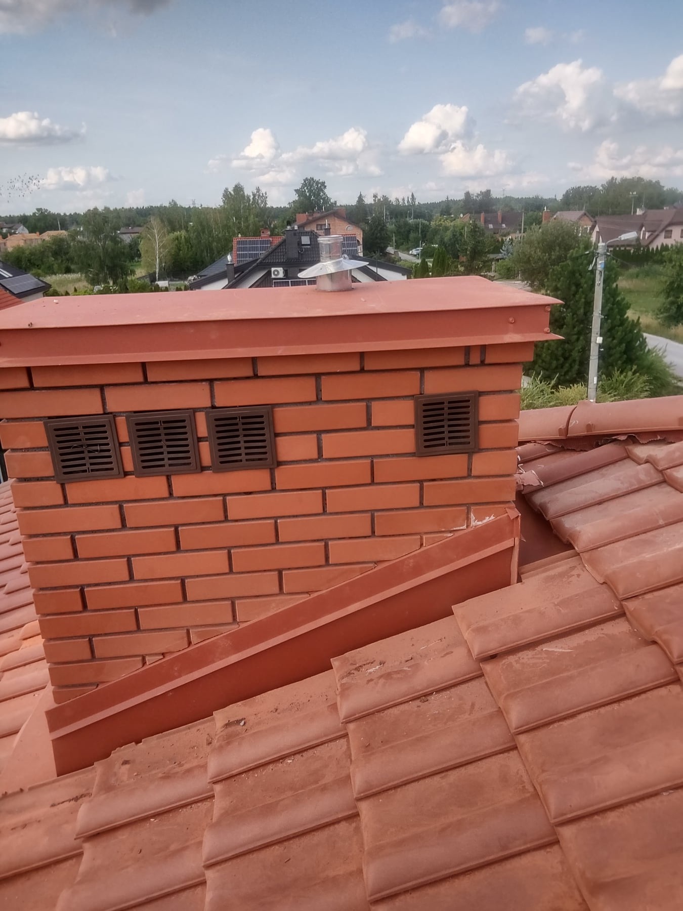 Czerwony ceglany komin z trzema kratkami wentylacyjnymi na dachu pokrytym czerwoną dachówką ceramiczną, widok na zabudowania w oddali pod błękitnym, pochmurnym niebem.