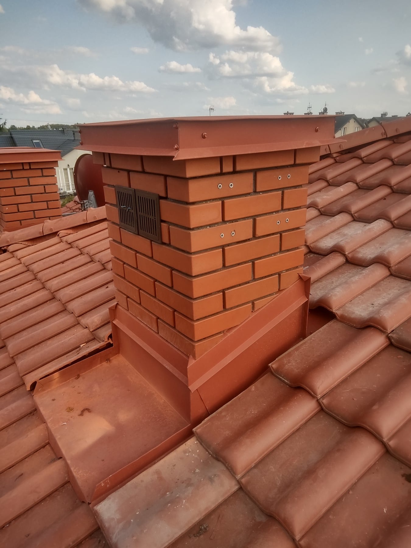 Ceglany komin z metalową obróbką na dachu z czerwonej dachówki, widoczne kratki wentylacyjne i mocowania, w tle zabudowa jednorodzinna i błękitne niebo z obłokami.