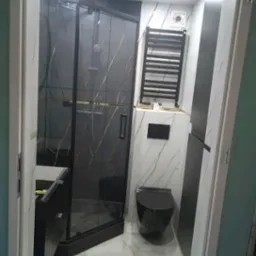 Nowoczesna łazienka z czarną kabiną prysznicową z marmurowym wzorem, wiszącą toaletą w czarnym kolorze i grzejnikiem drabinkowym na ścianie wyłożonej płytkami imitującymi marmur.