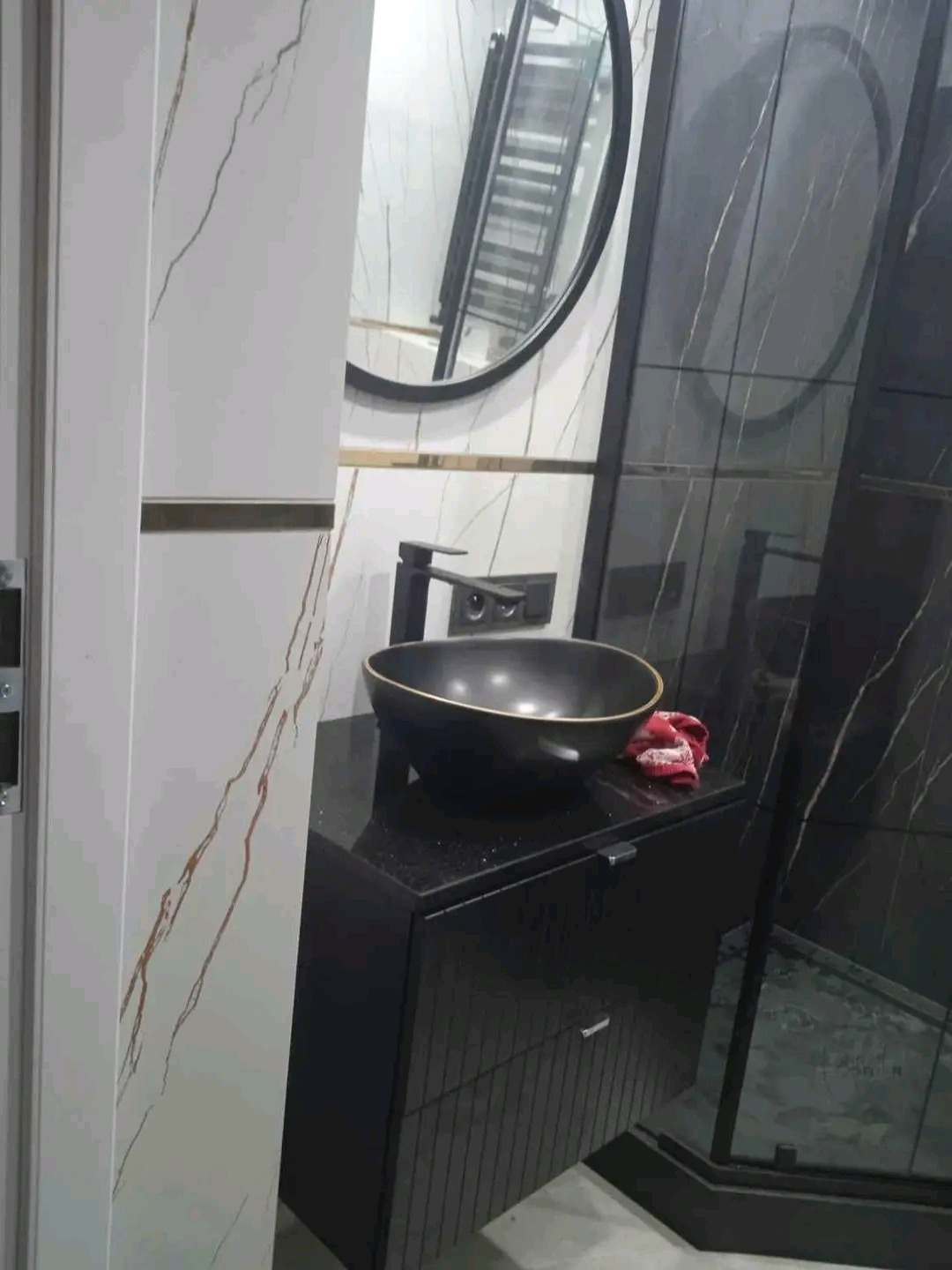 Czarna, okrągła umywalka nablatowa z baterią w kolorze czarnym, stojąca na czarnej szafce łazienkowej z szufladami, na tle białych i czarnych płytek imitujących marmur oraz okrągłego lustra.