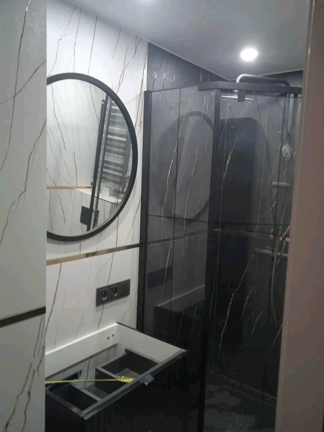 Nowoczesna łazienka z czarno-białymi marmurkowymi płytkami, okrągłym lustrem i czarną kabiną prysznicową z ciemnego szkła, widoczny fragment szafki pod umywalkę w trakcie montażu.