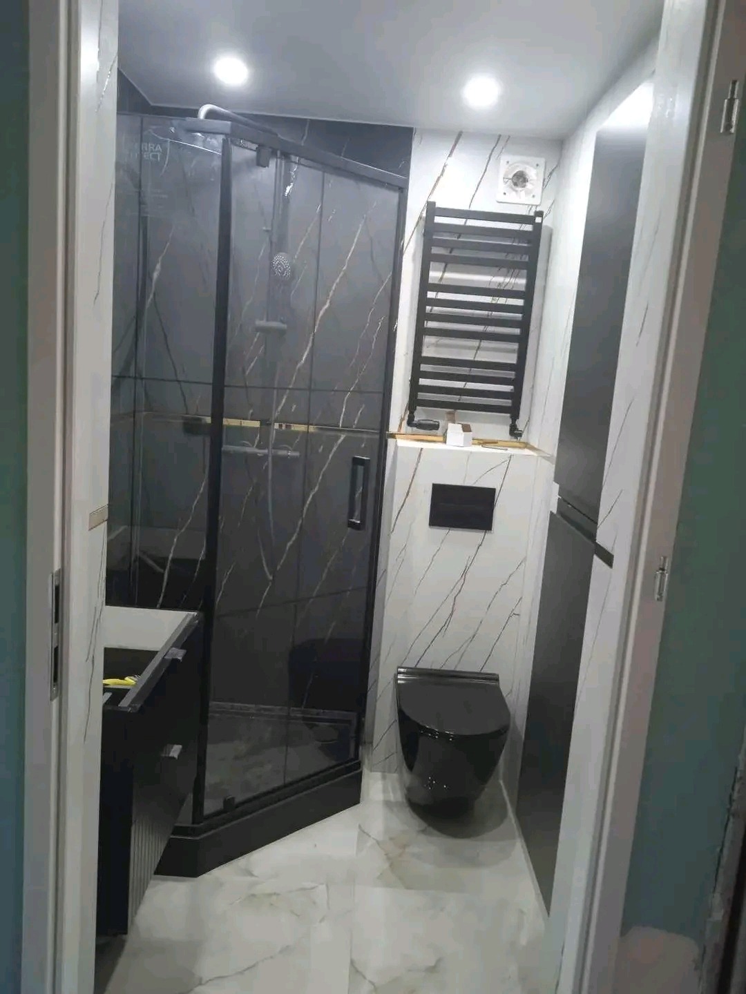 Nowoczesna łazienka z czarną kabiną prysznicową z marmurowym wzorem, wiszącą toaletą w czarnym kolorze i grzejnikiem drabinkowym na ścianie wyłożonej płytkami imitującymi marmur.