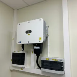 Biały falownik Huawei zamontowany na ścianie wewnątrz budynku, z widocznymi skrzynkami zabezpieczeń i przewodami.