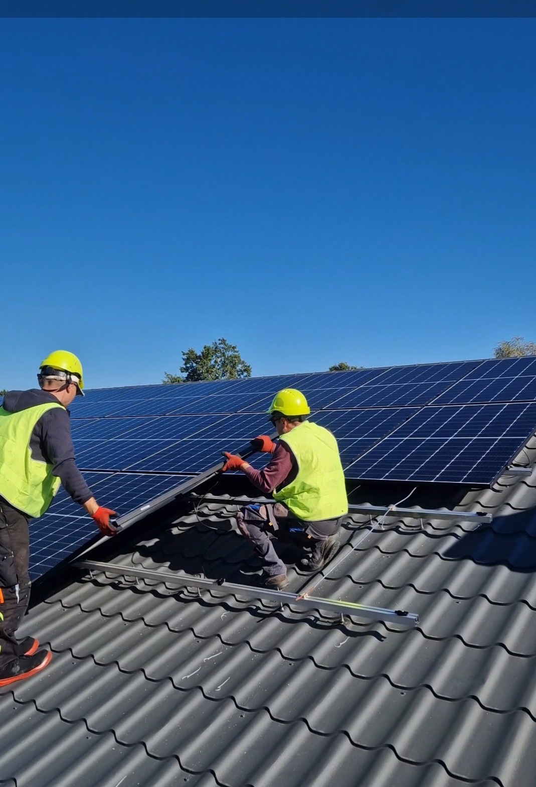 Montaż paneli słonecznych na dachu w Wilkowa Wsi przez dwóch instalatorów w kaskach i kamizelkach odblaskowych, widok z dołu na błękitne niebo.