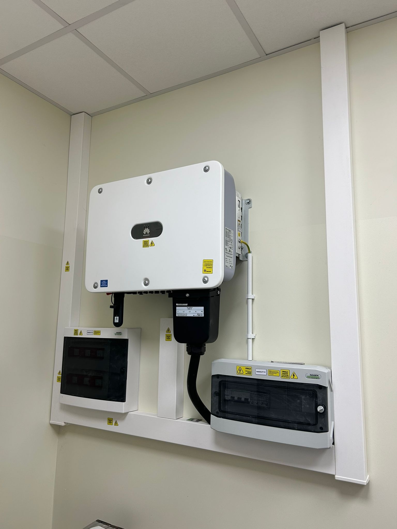 Biały falownik Huawei zamontowany na ścianie wewnątrz budynku, z widocznymi skrzynkami zabezpieczeń i przewodami.