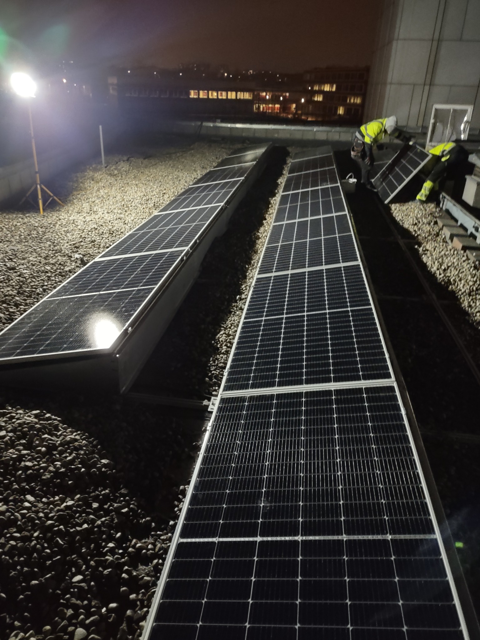 Montaż paneli słonecznych na dachu pokrytym żwirem, nocna sceneria oświetlona reflektorem, widoczni pracownicy w odblaskowych kamizelkach instalujący kolejne panele.