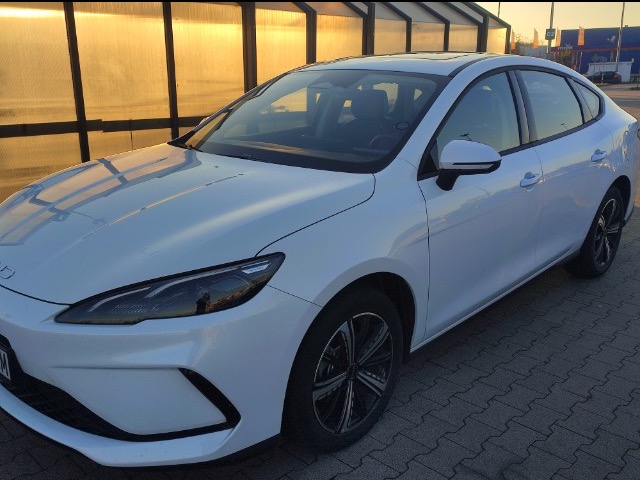 Biały, nowoczesny samochód typu sedan, zaparkowany na kostce brukowej, z widocznym fragmentem parkingu w tle i ciepłym, zachodzącym słońcem odbijającym się w karoserii.