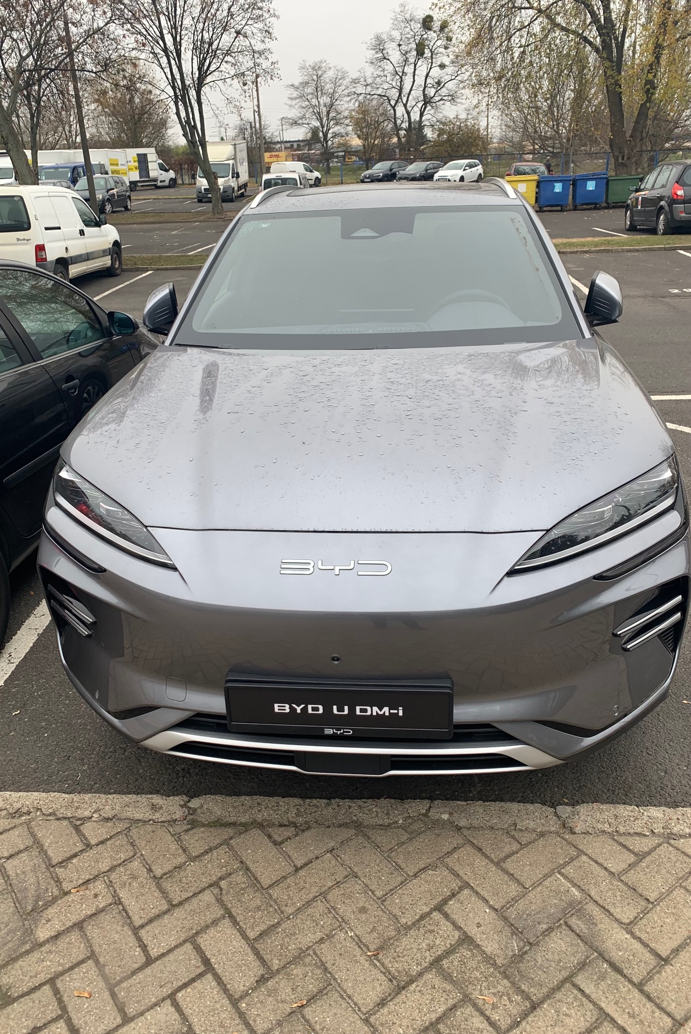 Szary samochód elektryczny BYD U DM-i na parkingu, widok z przodu, z kroplami deszczu na karoserii. Widoczne logo BYD i reflektory.
