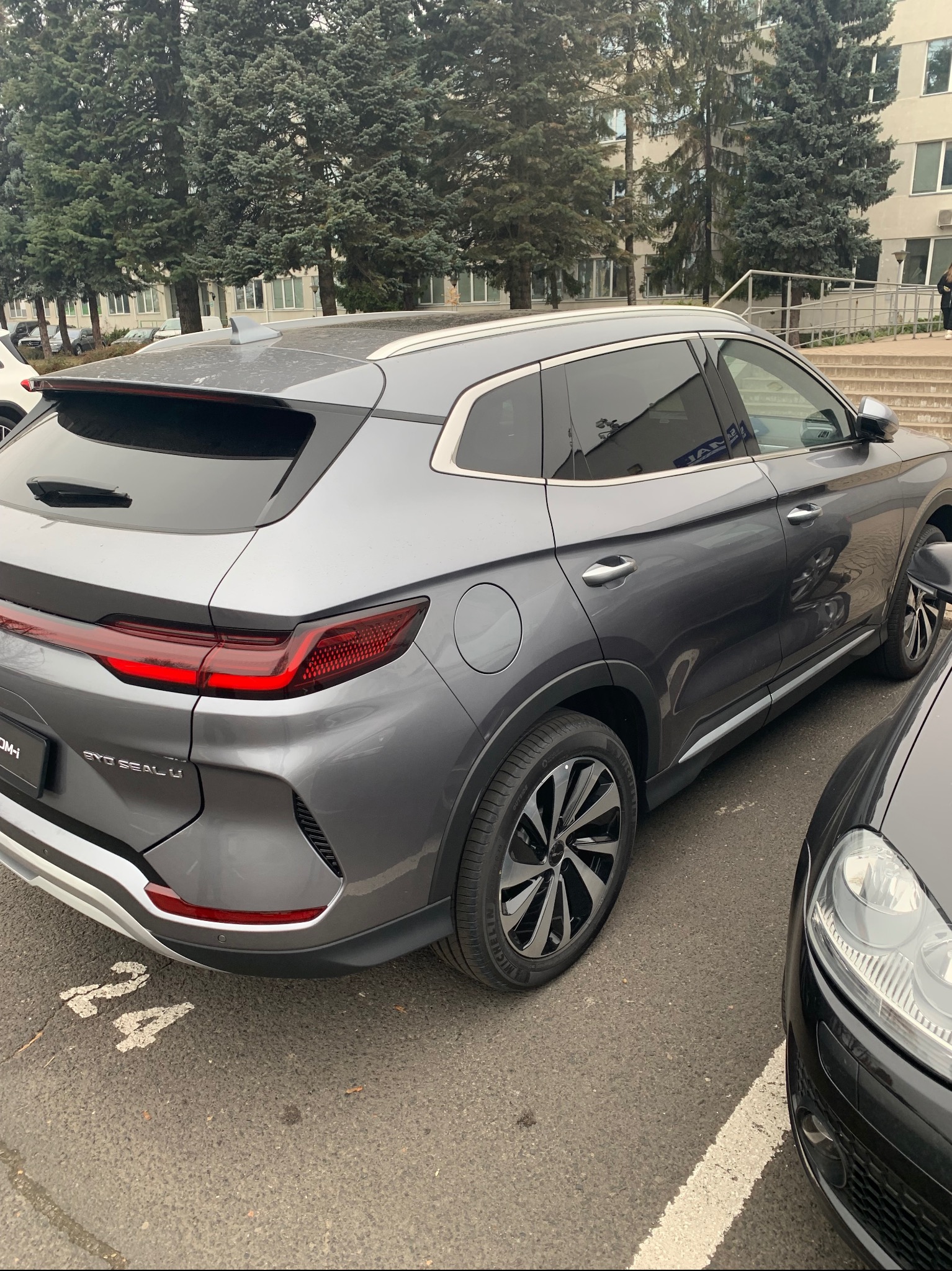 Szary SUV BYD Seal U zaparkowany na parkingu z numerem 24, widok z tyłu, z charakterystycznymi światłami LED i felgami. W tle budynek i drzewa.
