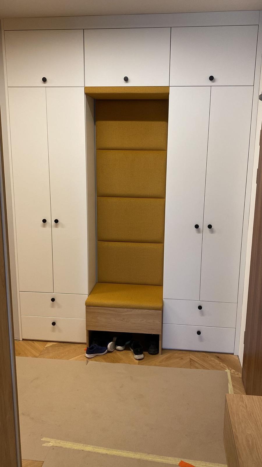 Biała szafa w przedpokoju z wnęką do siedzenia wyłożoną żółtą tapicerką, pod siedziskiem widoczne buty.